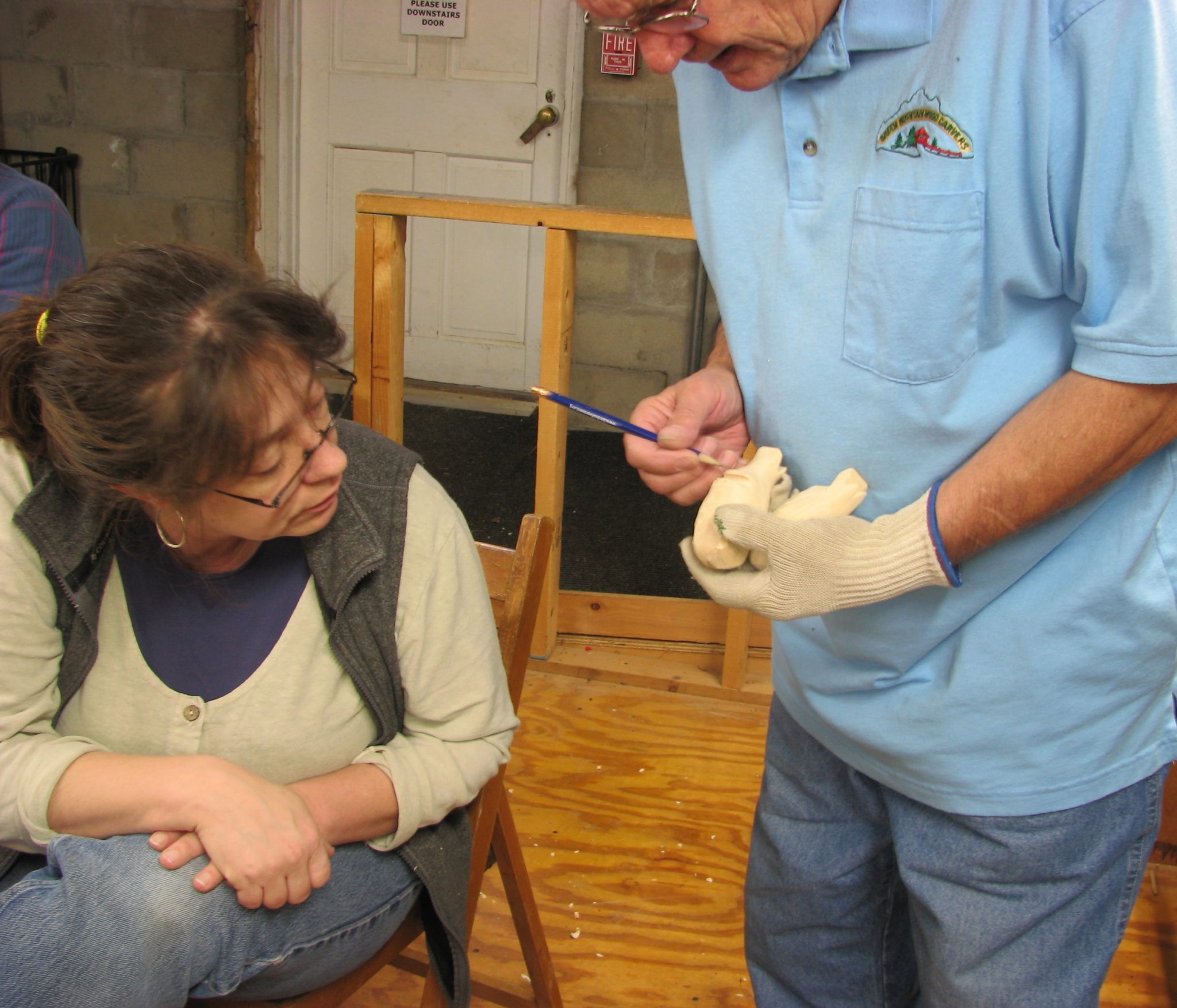 Senior man, standing, in blue shirt on right demonstrates woodcarving while adult woman, seated, looks on from left.