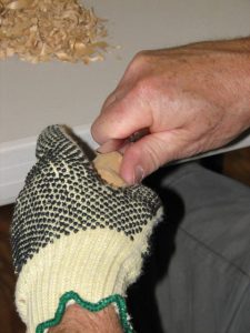 Hands of older, light-skinned person. The left hand is wearing a Kevlar carving glove and holding a partially-carved piece of wood. The right hand is holding a carving knife and is carving the wood.