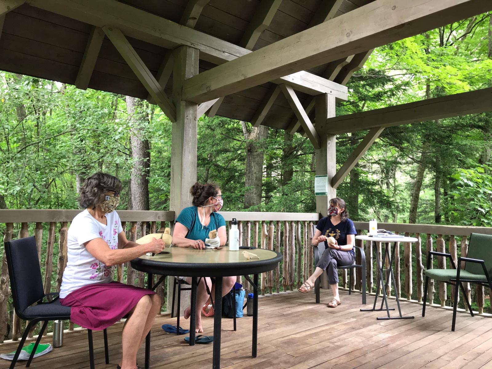 three women, wearing fabric masks and physically distancing, converse while carving birds from wood at the Birds of Vermont Museum's walk-in treehouse