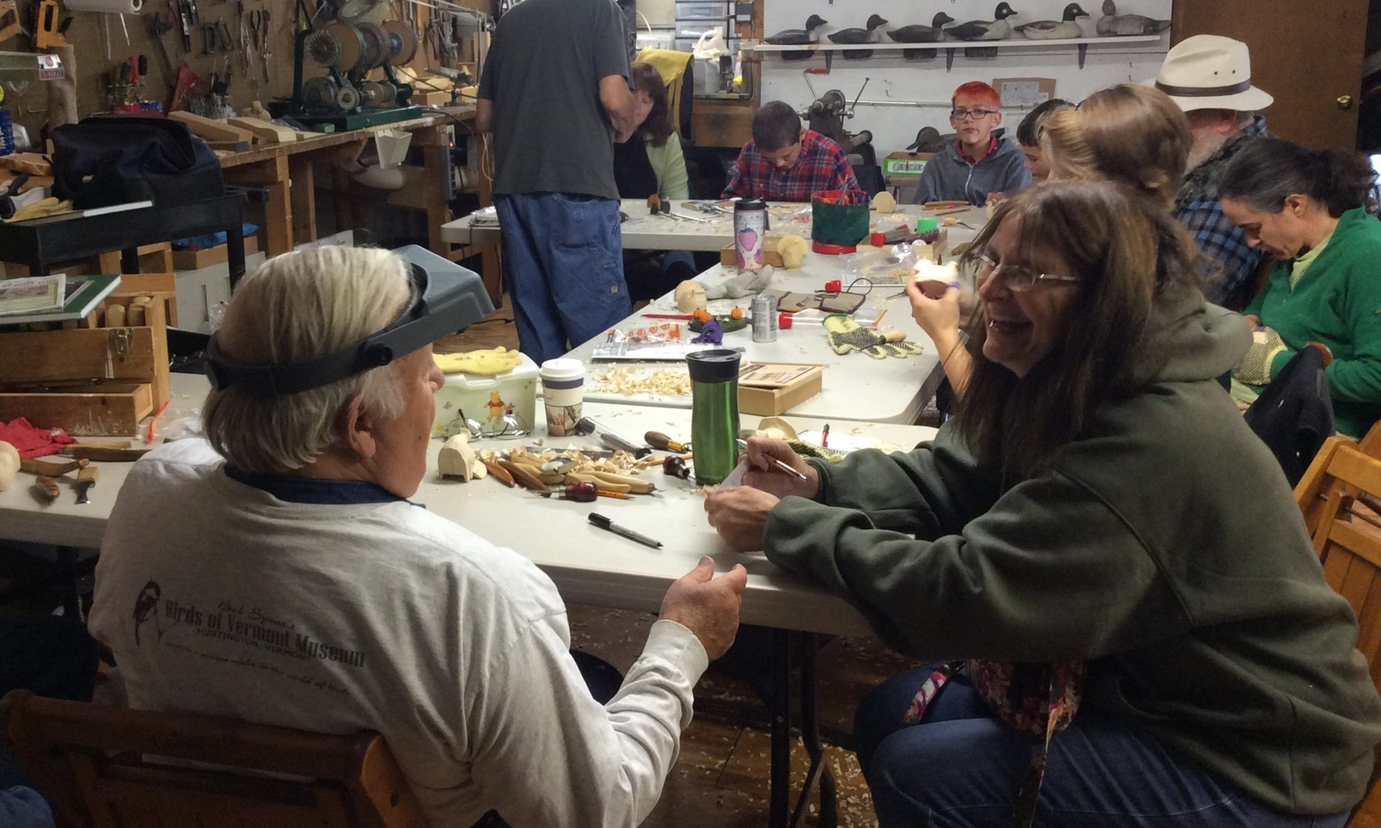 Several woodcarvers carving, painting, and chatting at tables in the workshop at the Birds of Vermont Museum.