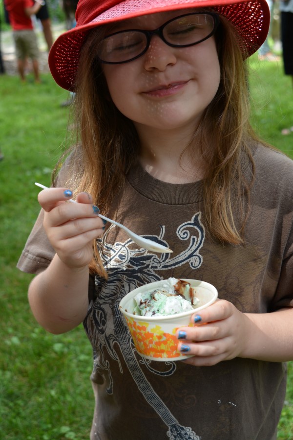 Child in glasses grins at camera with eyes shut while holding a paper cup with ice cream.
