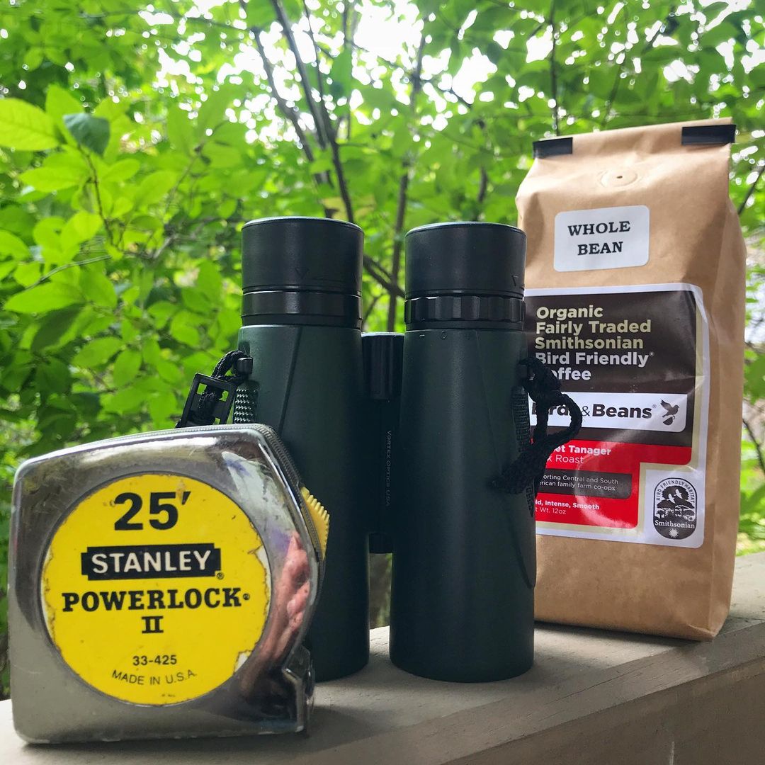 A Stanley brand 25' metal measuring tape; a pair of black binoculars; a bag of Birds and Beans coffee (scarlet tanager dark roast). All three item are line d up on a wooden railing, with green foliage behind them.