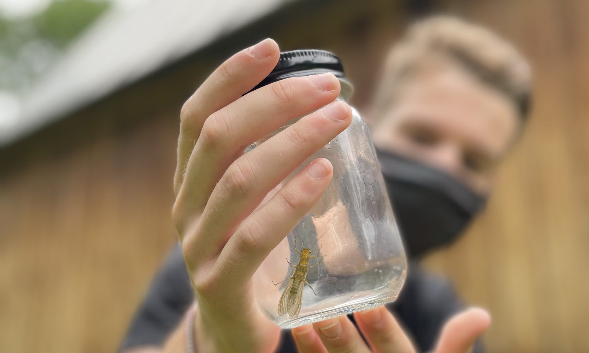 A stonefly in a glass jar is held toward the camera by a young white man
