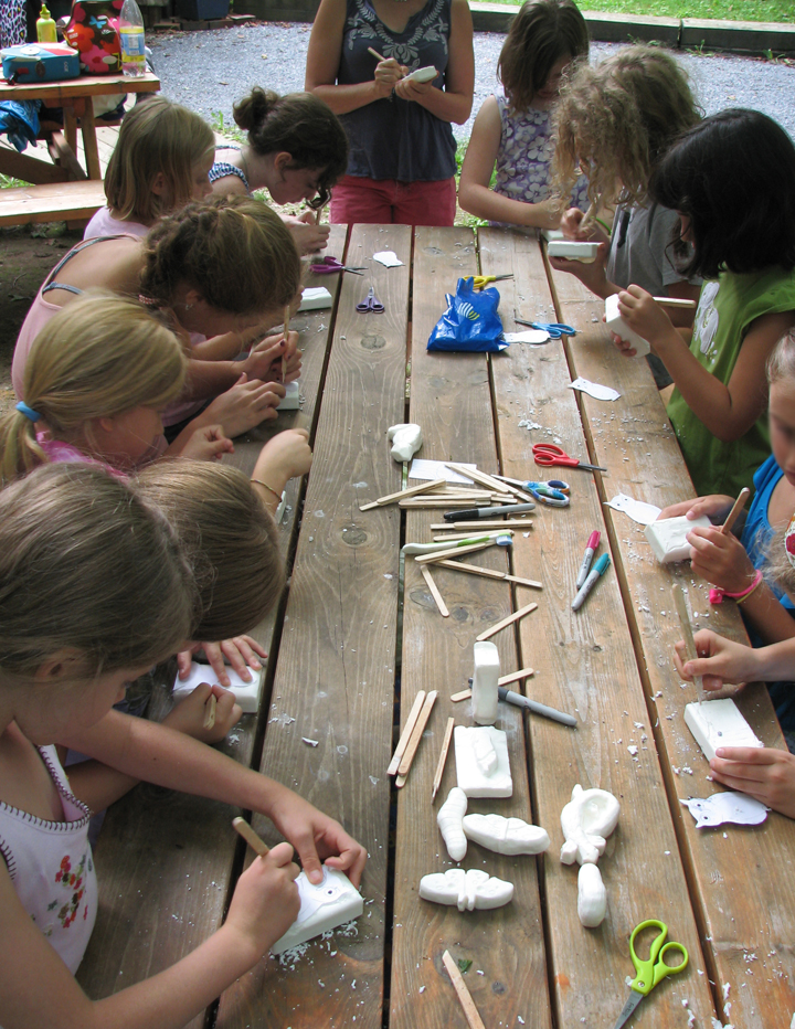several children around a wooden table carving white bars soap using tools made of craft sticks