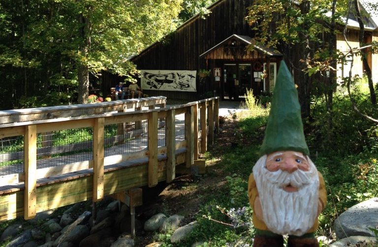 small wooden gnome, carved by David Tuttle, in foreground, with railing of Museum's wooden bridge and the Museum entrance visible in background.