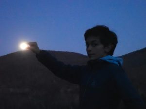 A young person glances at the photographerwhile appearing to hold the full moon between their thumb and index finger. The moon is rising above curved hills in the background. The sky is dark blue and the person and hills are almost silhouetted.
