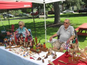 two older people sitting behind a table wit hmany small woodcarvings on it