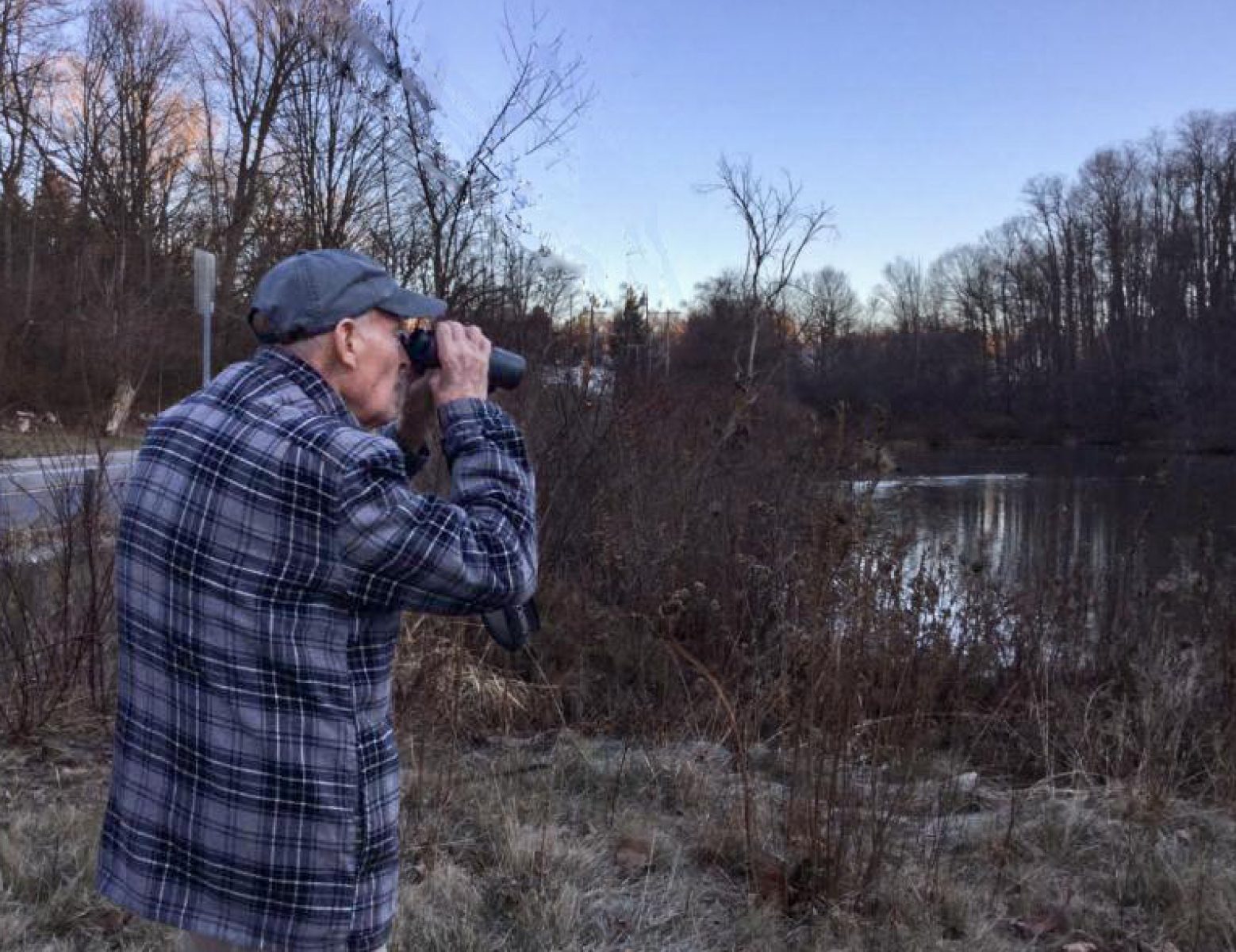 an elderly man in a blue plaid shirt looks away from the photographer, through binoculars, at a late-winter pond and trees