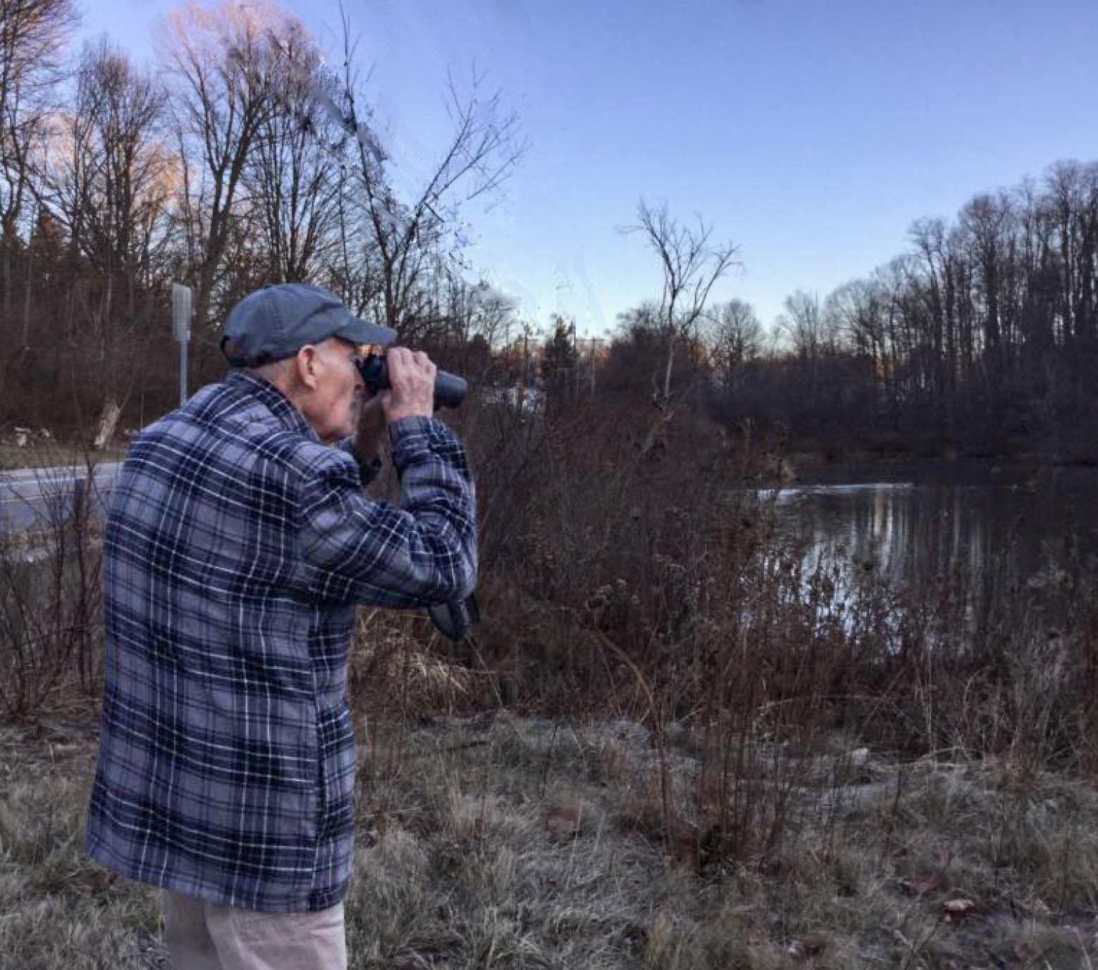 an elderly man in a blue plaid shirt looks away from the photographer, through binoculars, at a late-winter pond and trees
