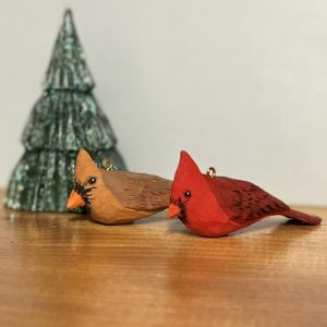 Three painted woodcarvings: a male Northern Cardinal, a female Northern Cardinal, and an evergreen tree. They sit on a wooden surface against a pale background, with the male in front and the tree in back, overlapping. The birds are ornaments: they have small brass loops on their backs through which a cord could be threaded. The tree is a somewhat layered cone in dark green with some white and glitter on some of the ridges of its branches.