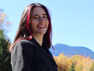 Rachel Mirus: A light-skinned woman with straight, long brown and red hair, smiles in three-quarter view at camera. She is visible from shoulders up. She is outdoors with golden autumn foliage in background and a blue mountain ridge behind. Image shown with permission.
