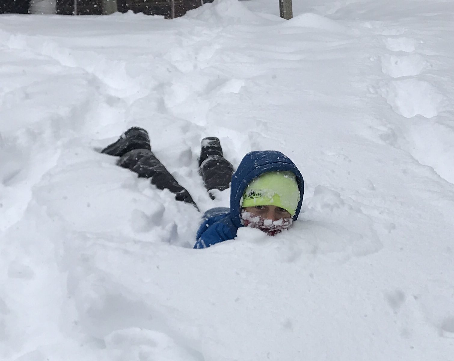 Child playing in deep snow. Child is wearing green hat,blue hooded parka, black snow pants, but because is prine, kid is partially hidden by snow.