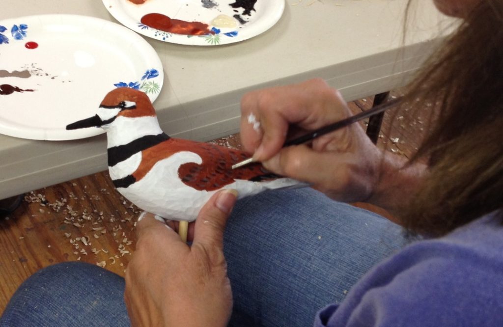 A wooden killdeer being held in one hand while being painted . View is over the painter's left shoulder.