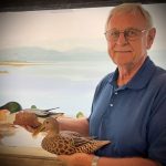 An older white man (Bill Paterson) wearing glasses and a blue shirt holds two woodcarvings (Northern Shoveler male and female) in front of a painting of Lake Champing and the Adirondacks.