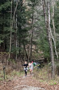 Three runners coming uphill toward the viewer. They are on a gravel trail rising from a creek, with evergreens and other trees behind them and to their left and right. Other tall, autumn-dried plants are on the sides of the trails.