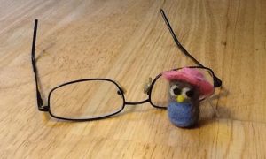 Mini needle-felted owl (gray with blue belly and pink hat) in from of black wire-framed glasses