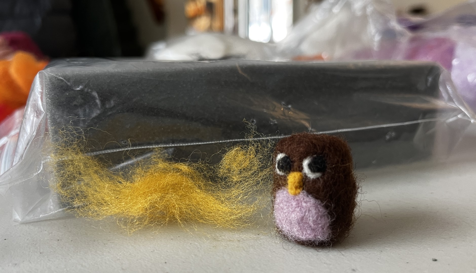 brown mini owl and yellow roving, in front of a plastic-wrapped block of felting foam