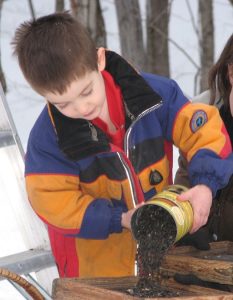 A young child in a multi-colored winter coat pours black oil seed into a tray.