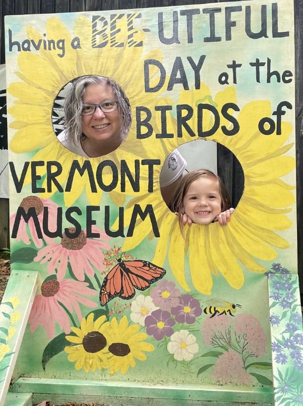 An older person and younger person smile through circular openings in a large painted signboard. The board says 'Having a BEE-utiful Day at the Birds of Vermond Museum'. The openings are the centers of two big yellow flowers; other flowers, a monarch butterfly, and a honeybee are painted on the board.