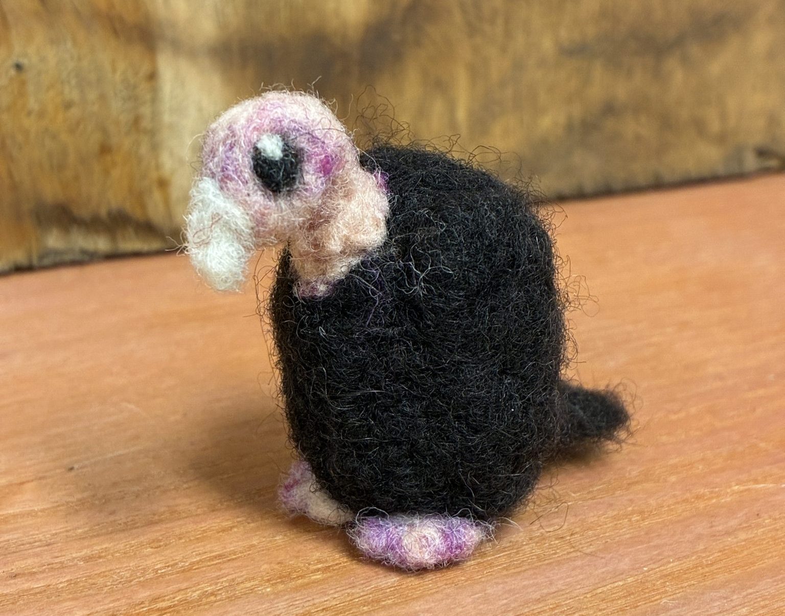 A turkey vulture made of needle-felted wool stands on a wooden surface.