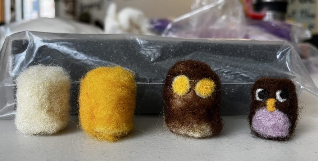 a sequence of 4 needle felted mini-owls in progress, from the core to a color applied to one with eyes, to a finished owl. Each is roughly cylindrical and about 1" tall.