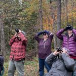 6 humans in brightly colored coats sit and stand and look through binoculars. Behind them are autumn deciduous trees with some brightly colored leaves still visible. 