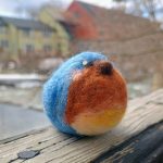 Round cozy needle-felted male Eastern Bluebird. He sits on a faded wooden rail, with blurred farm buildings behind him. He appears to be looking up and to the right, in 3/4 view. [Made by Grace Safford; photo used with permission.]