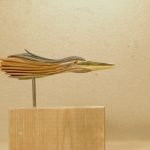 Woodcarving Sculpture: a head and upper neck of of a heron reaches horizontally. The wood is tinted in semi-transparent blues and reddish colors; the beak is gray and yellow. The sculpture stands on a thin post and a wooden block.