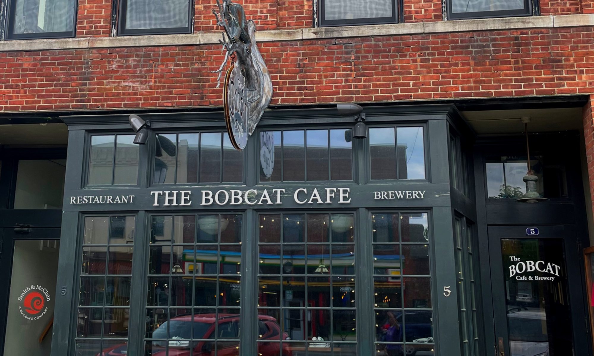 The Bobcat café building front: Several large mullioned windows with dark green trim on the first story; brick facing on the upper story. The door to the café is on the right.