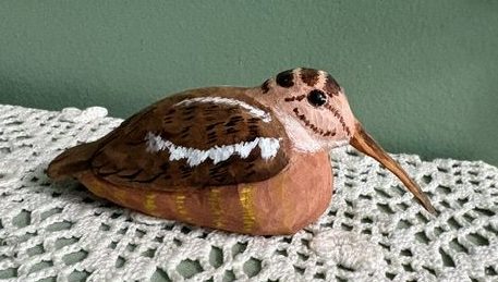 Roosting Woodcock (Scolopax minor) carved of wood and painted to suggest live birds' plumage. This carving appears to be roosting (sitting on the ground). It has a long beak, a pale brown head with a darker brown eye-line and chin line and dark patches across its crown. It has darker brown wings with patchy white wing bars and even darker brown flecks. Its breast and belly are a medium brown with slightly yellow patches.