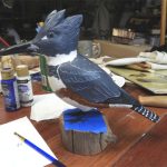 Belted Kingfisher, female, carved of wood and painted. Nearly life-size. It sits on a block of wood on a work table.