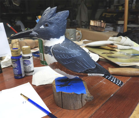 Belted Kingfisher, female, carved of wood and painted. Nearly life-size. It sits on a block of wood on a work table.