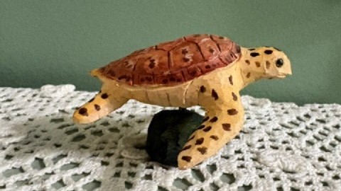 Small sea turtle woodcarving in profile. Brownish and light brown turtle is on a black stand which rests on a lace-covered table surface. Carving and photo by David Tuttle.